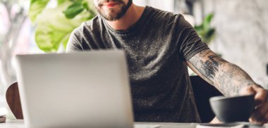 Yakışıklı sakallı hippi, kafeteryasında kahve olan dizüstü bilgisayarı kullanır ve ona bakar. İletişim ve teknoloji kavramı.