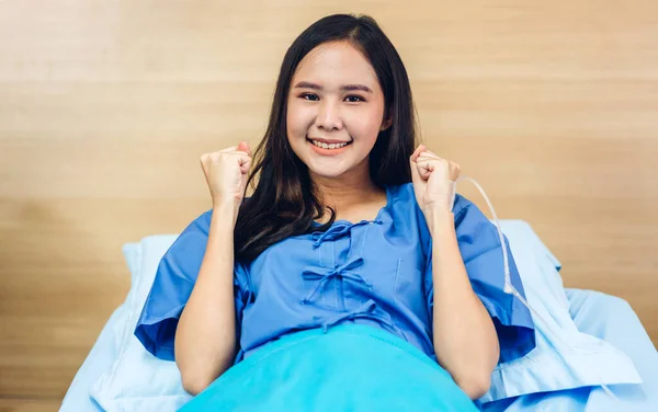 Portrait of smiling asian woman patient sitting on bed looking at camera raising her fist with health medical care express trust and Insurance concept in room at hospital