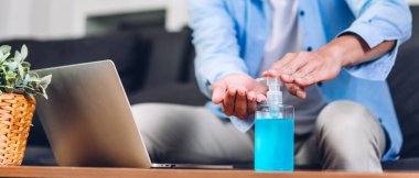 Koronavirüs için karantinada alkol jeliyle ellerini yıkayan genç bir adam sosyal mesafeli koruyucu maske takıyor ve evde çalışan dizüstü bilgisayarı kullanıyor. Evden çalışma ve covid19 konsepti