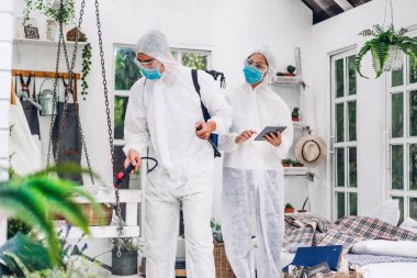 Koruyucu maske takan dezenfekte işçileri ve müşteri evindeki Coronavirüs 'ü öldürmek için beyaz takım elbise dezenfektan sprey temizlik virüsü kullanan profesyonel ekipler.
