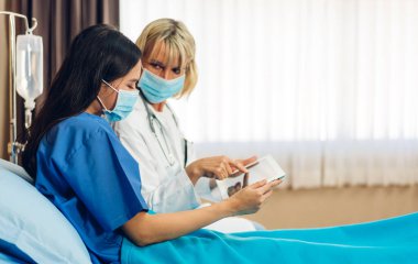 Steteskopla koruyucu maske takan kıdemli kadın doktor, hasta kadın hastalarla hastanede tabletle yapılan kontroller hakkında konuşmak ve danışmanlık desteği sağlıyor.