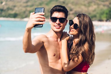 Romantik aşıklar genç çift tropik sahilde birlikte dinleniyor. Erkek ve kadın akıllı telefonlarıyla selfie çekiyor ve hayatın tadını çıkarıyor. Yaz tatilleri.