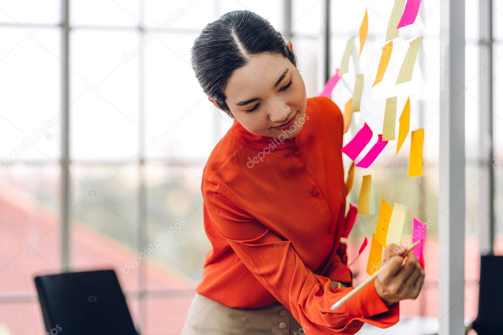 Casual creative happy smile asian business woman planning strategy analysis putting post it stickers note to share idea for startup project on glass board at modern office