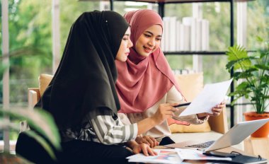 Genç, gülümseyen mutlu Asyalı Müslüman kadın evde dizüstü bilgisayar, tablet çalışma ve video konferans kullanarak rahatlıyor. Ev konseptinden iş.