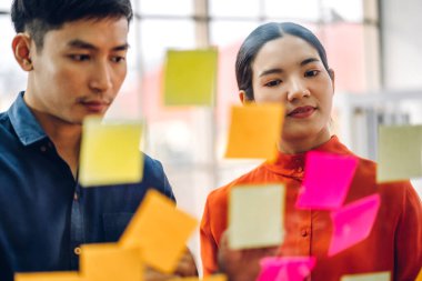 Gündelik yaratıcı mutlu iki gülümseme Asyalı iş adamları strateji analizi ve beyin fırtınası planlıyorlar. Çıkartma yapıştırmaları yapıştırıyorlar ve modern ofisin cam tahtasında başlangıç projesi için fikir paylaşıyorlar.