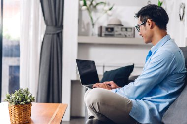 Genç yaratıcı gülümseyen mutlu Asyalı adam masaüstü bilgisayarını kullanarak rahatlıyor ve ev konseptinden online sohbet yapıyor.