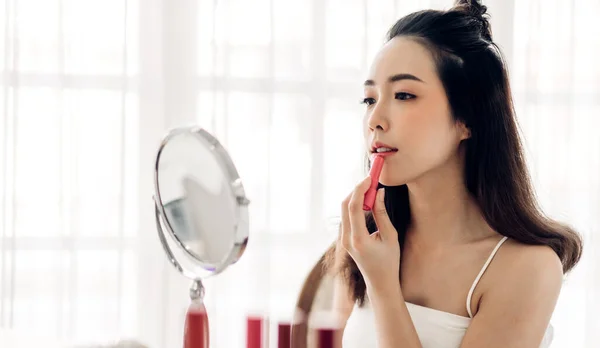 Smiling beautiful asian woman fresh healthy skin looking on mirror and enjoying applying lips with red lipstick at home.facial beauty and cosmetic concept