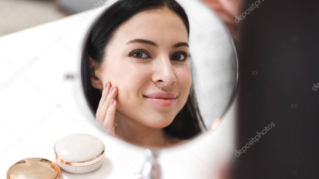 Smiling of young beautiful pretty woman clean fresh healthy white skin looking at mirror.girl touching on her face with hand and applying cream at home.spa and beauty concept