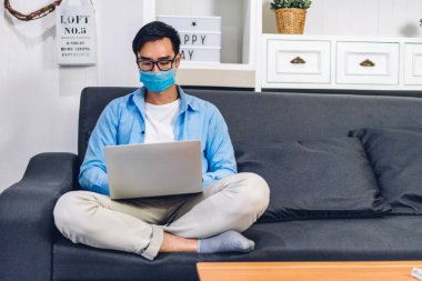 Genç Asyalı adam ev konseptinden kalma koruyucu maske takarak karantinada bilgisayar kullanıyor ve video konferansı yapıyor.