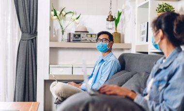 Genç Asyalı çift dizüstü bilgisayar kullanıyor ve Coronavirus için karantinada görüntülü görüşme yapıyor. Ev konseptinden sosyal mesafeli koruyucu maske takıyorlar.