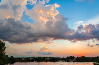 Mavi ve pembe tonlarda akşam gökyüzü, kıvırcık bulutlar ile, uzak bir ormanlık kıyı ve kentsel binalar ile gölün yüzeyi üzerinde batan güneş tarafından aydınlatılmış.