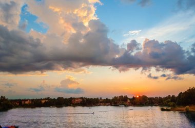 Mavi ve pembe tonlarda akşam gökyüzü, kıvırcık bulutlar ile, uzak bir ormanlık kıyı ve kentsel binalar ile gölün yüzeyi üzerinde batan güneş tarafından aydınlatılmış.