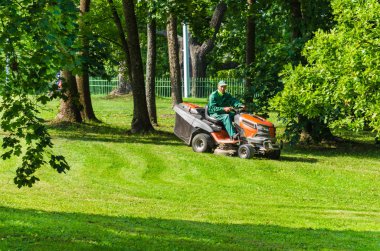 Gatchina, Leningrad bölgesi, Rusya - 30 Ağustos 2019: yeşil tulumlu işçi bir yaz parkında çim biçme makinesiyle çimleri biçiyor.