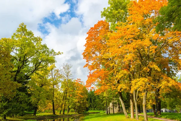 Park 'ta sonbahar manzarası. Taş piramidi, sarı, yeşil, kırmızı yapraklar çimlerin üzerinde, mavi gökyüzünde bulutlarla.
