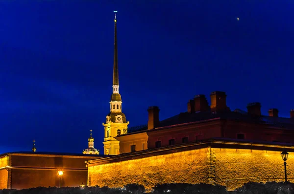 The wall of an ancient fortress on the river bank with a high tower and spire, at night, illuminated by the light of lanterns and searchlights against the dark sky.