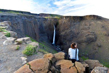 Young woman is sitting at the rock in front of spectacular view at Litlanesfoss waterfall surrounded with vertical basalt columns, Fljotsdalshreppur municipality of Eastern Iceland  clipart