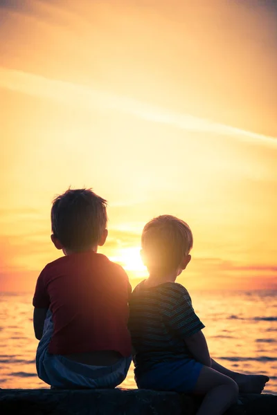 Two boys sitting on the rock at the beach at sunset — Stock Photo, Image