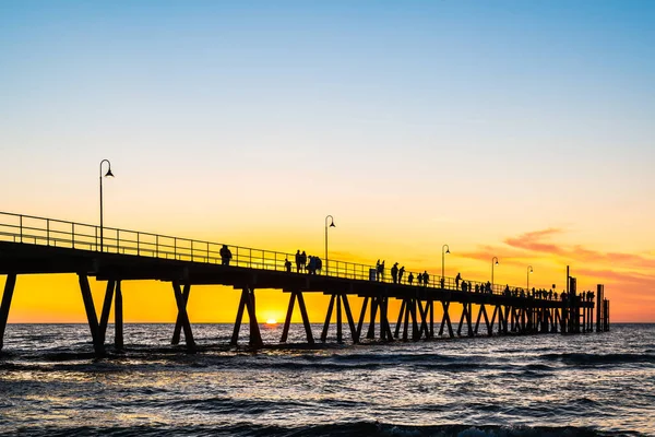 Stunning sunset at Glenelg Beach jetty, with silhouettes of people leisurely strolling along the pier in Adelaide, South Australia