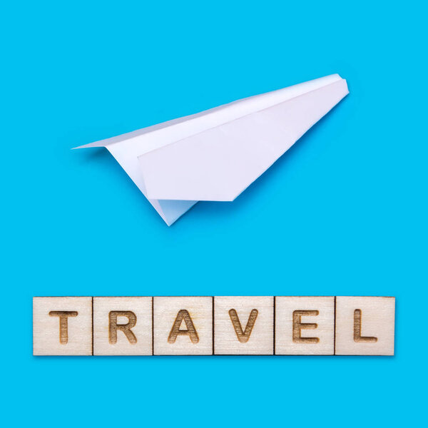 Concept on the theme of travel. White origami plane on a blue background. The word Travel is laid out of wooden blocks.