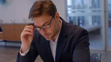 Takım elbiseli orta yaşlı bir işadamı ofis masasında dudaklarının yanında bardak tutarak oturuyor. Sahne konsantrasyon, iş planlaması, ciddi karar verme ve profesyonel kurumsal atmosfer gösteriyor.