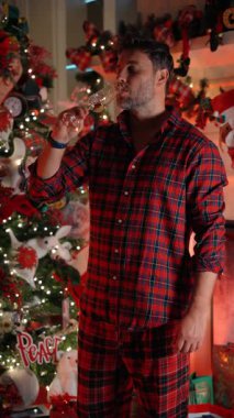 Kırmızı ekose pijamalı bir adam süslü Noel ağacının yanında durmuş evde şampanya içiyor. Sıcak tatil havası, sıcak ışıklar, şenlikli kutlamalar, aile neşesi ile yeni yıl ve Noel gelenekleri.