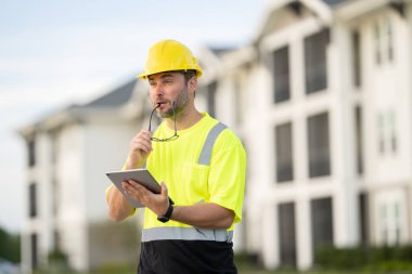 Sarı güvenlik yeleği giymiş bir adam kasketli bir şekilde tablete bakıyor. Cihaza odaklanmış durumda, muhtemelen güvenlik bilgileri ya da talimatları kontrol ediyor.
