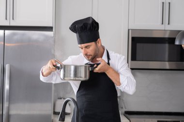Profesyonel üniformalı modern şef. Kaşıkla tencereye çorba karıştıran erkek aşçının eline yakın plan. Yemek pişirme, aşçılık ve insan konsepti - erkek aşçı tencereli veya tencereli.