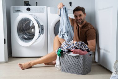 Çamaşır odasında çamaşır makinesinin yanında giysileri sıralayan mutlu bir adam. Adam kirli çamaşırlarıyla çamaşır makinesinin yanında bir evin zemininde oturuyor. Bir adam çamaşırlarını evde yıkıyor. Kirli çamaşırları olan bir adam.