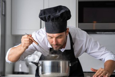 Profesyonel üniformalı modern şef. Kaşıkla tencereye çorba karıştıran erkek aşçının eline yakın plan. Yemek pişirme, aşçılık ve insan konsepti - erkek aşçı tencereli veya tencereli.
