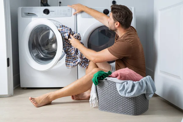 Çamaşır odasında çamaşır makinesinin yanında giysileri sıralayan mutlu bir adam. Adam kirli çamaşırlarıyla çamaşır makinesinin yanında bir evin zemininde oturuyor. Bir adam çamaşırlarını evde yıkıyor. Kirli çamaşırları olan bir adam.