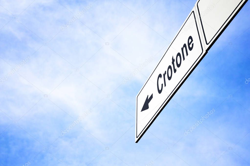 White signboard with an arrow pointing left towards Crotone, Calabria, Italy, against a hazy blue sky in a concept of travel, navigation and direction. Path included for the signboard