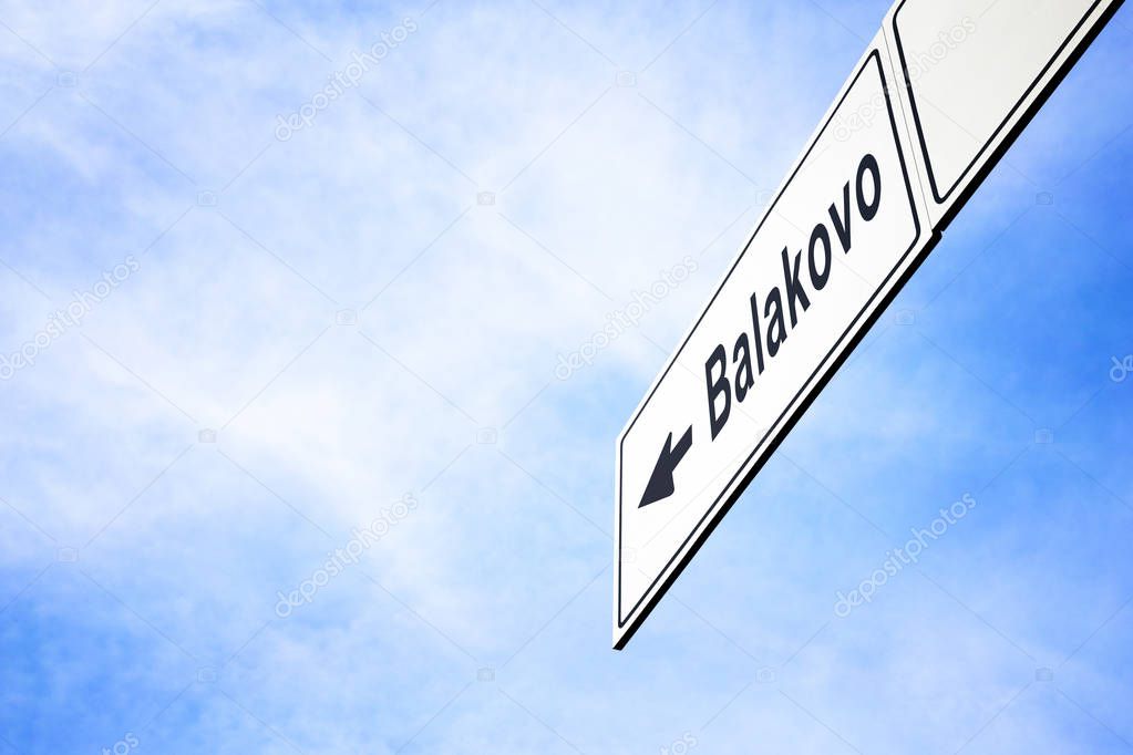 White signboard with an arrow pointing left towards Balakovo, Saratov Oblast, Russia, against a hazy blue sky in a concept of travel, navigation and direction. Path included