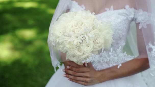 Jovem noiva bonita em vestido branco de casamento com buquê de rosas brancas que ficam em um parque no dia de verão — Vídeo de Stock