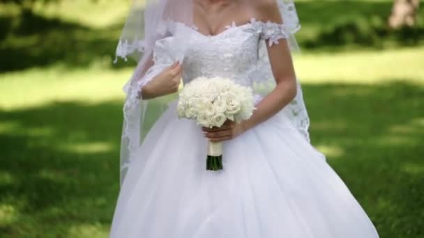 Jonge mooie bruid in bruiloft witte jurk met boeket witte rozen verblijf in een park in de zomerdag — Stockvideo
