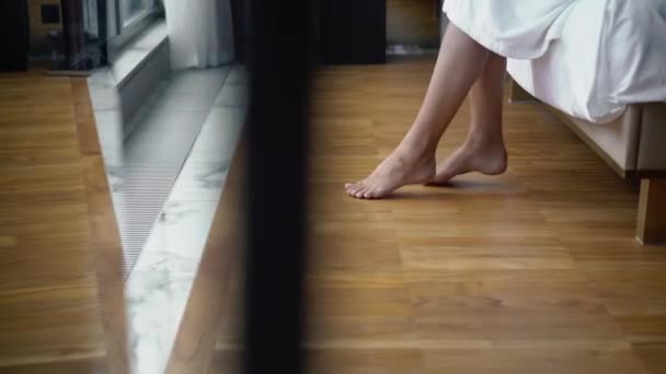Jonge vrouw in badjas zittend op een bed in de slaapkamer blote voeten in hotel — Stockvideo