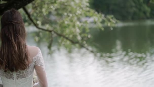 La sposa con il bouquet in mano è in piedi sullo sfondo di un fiume. Vista posteriore. Nastri su mazzo che saluta da vento . — Video Stock