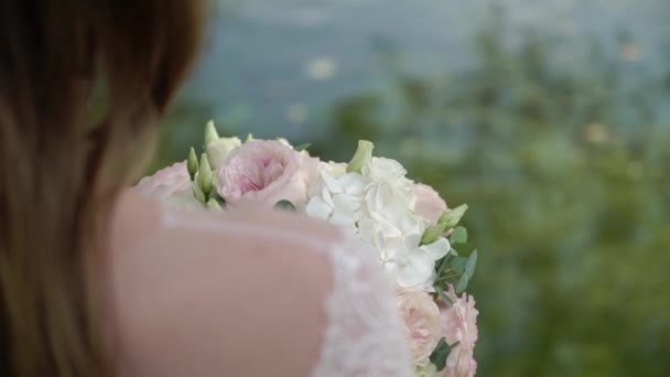 The bride with bouquet in her hands is standing against the background of a river. Rear view. Ribbons on bouquet waving from wind. — Stock Video