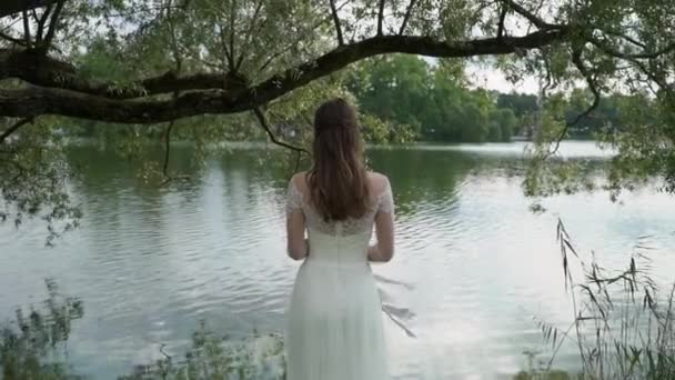 La novia con el ramo en las manos está de pie sobre el fondo de un río. Vista trasera. Cintas en ramo saludando del viento . — Vídeos de Stock