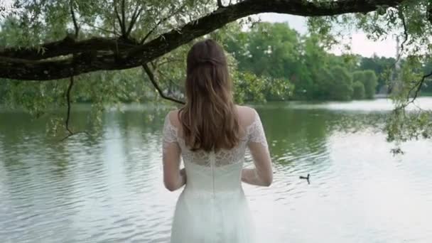 Nevěsty s kyticí v ruce stojí na pozadí řeka. Zadní pohled. Stužky na kytice mávat z větru. — Stock video