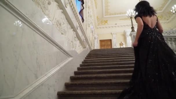 Young sexy brunette woman in black evening dress going upstairs at the marble stone ladder in palace or hotel — Stock Video