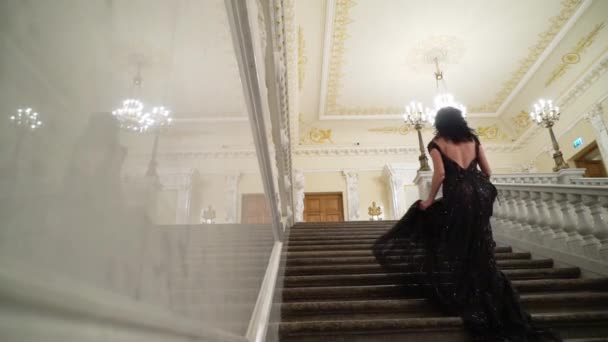 Giovane donna bruna sexy in abito da sera nero andando al piano di sopra alla scala in pietra di marmo a palazzo o in hotel — Video Stock