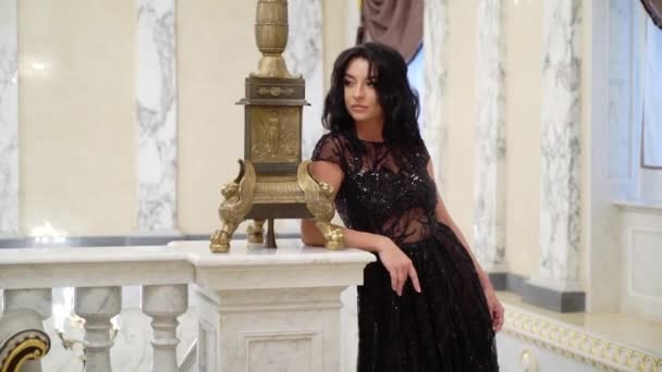 Young sexy brunette woman in black evening dress posing at the marble stone ladder in palace or hotel — Stock Video