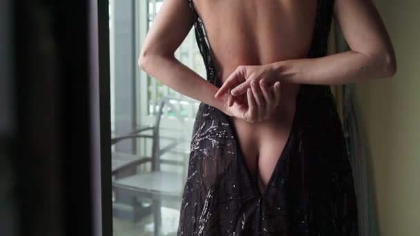 Junge sexy brünette Frau in schwarzem Luxuskleid übernachtet im Schlafzimmer in der Nähe des Fensters und zieht sich aus. — Stockvideo
