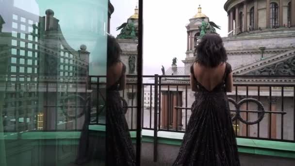 Молодая сексуальная брюнетка в черном роскошном платье, стоящая на балконе в городе — стоковое видео