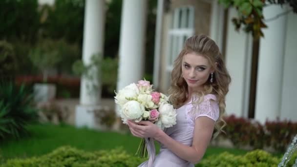 Молодая блондинка в нижнем белье позирует со свадебным букетом в саду возле фонтана — стоковое видео