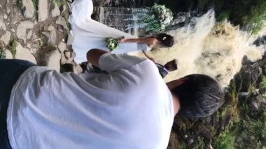 Dikey video. Genç düğün çifti töreninde şelale yanında kalıyorum. Adam alarak fotoğraf ve video. Fotoğrafçı sahne arkasında