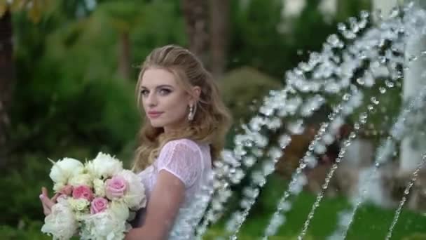 Молода блондинка в нижній білизні позує з весільним букетом в саду біля фонтану — стокове відео