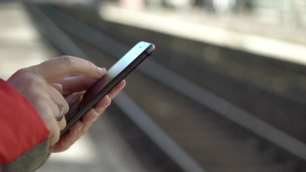 Молода жінка стоїть на стаціонарній платформі, чекаючи на поїзд зі смартфоном — стокове відео