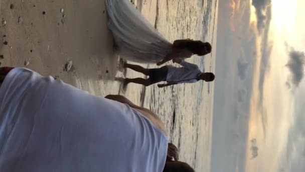 Verticale video. Fotografen het nemen van foto's van de bruid en bruidegom wandelen op het strand in de buurt van zee. Vakantie bruiloft foto sessie bij zonsondergang — Stockvideo