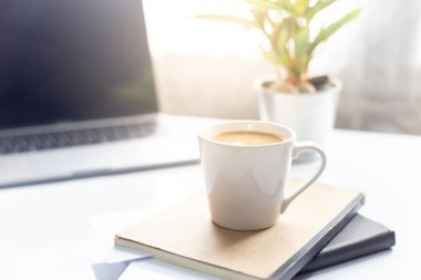 Sabah güneş ışığında dizüstü bilgisayarlı kahve fincanı, arka planda çalışma alanı.